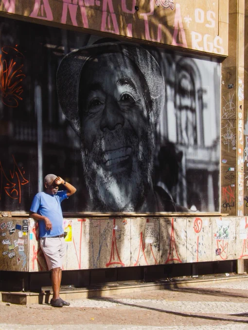 A person stands next to a large outdoor photograph of a smiling elder displayed on a wall covered with graffiti.