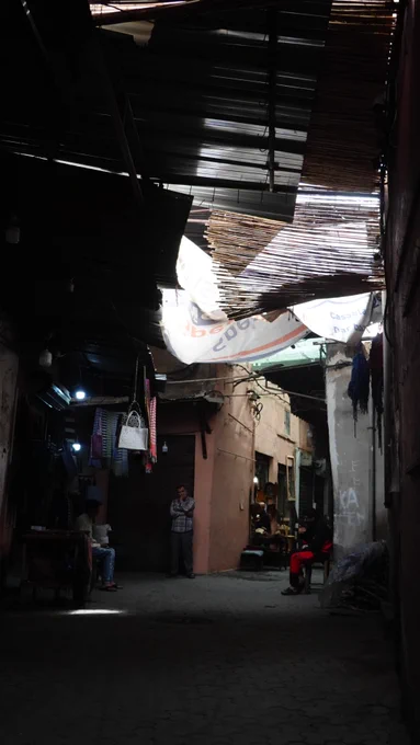 A narrow, dimly lit alleyway in a market, with partially visible banners and people engaged in everyday activities.