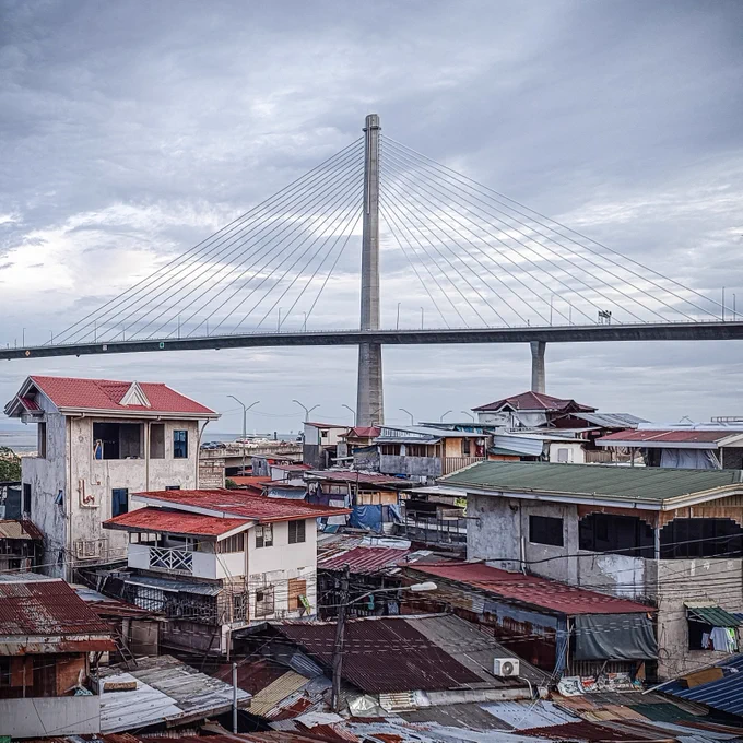 View of a riverside community with traditional houses in the foreground and the modern Expressway bridge spanning the river in the background under a cloudy sky.