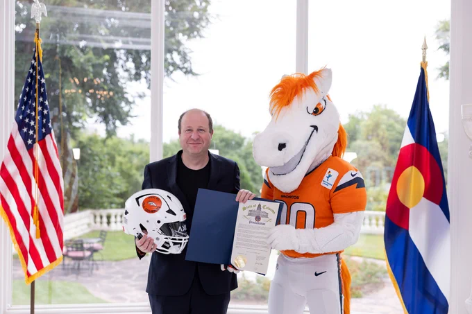 Colorado Governor Jared Polis and the Denver Broncos mascot pose with a proclamation and helmet between U.S. and Colorado flags.