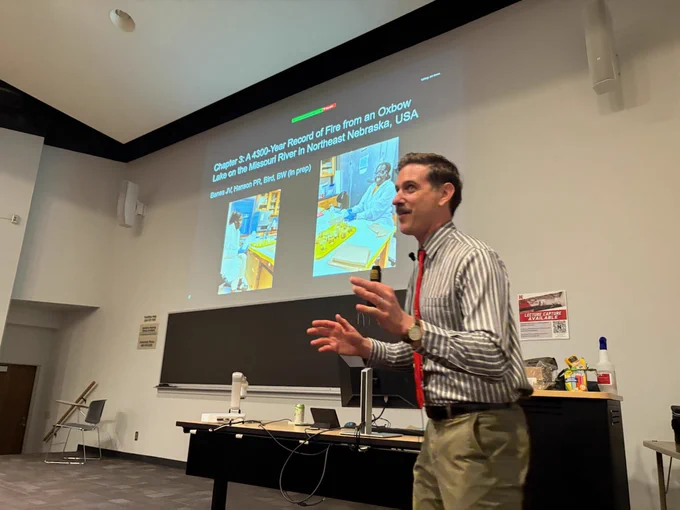 a young male presenter enthusiastically speaks to a crowd at the front of a university lecture hall