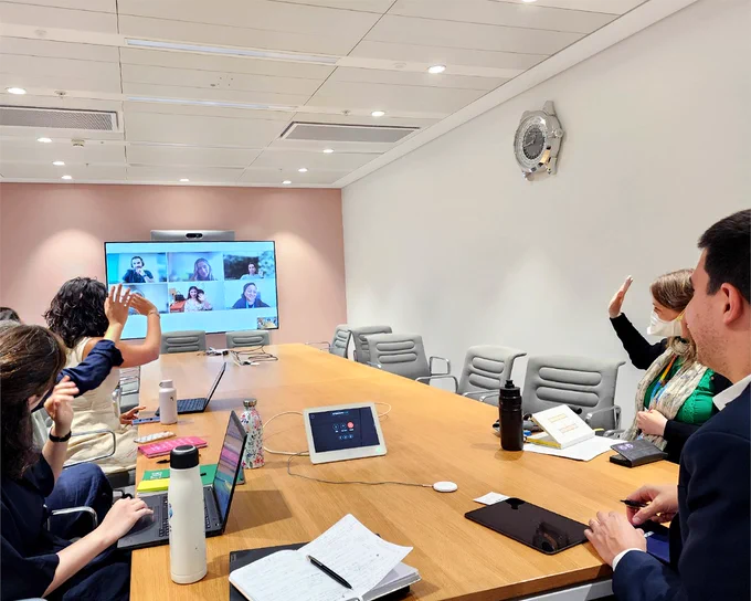 A conference room with five people seated around a table, participating in a video conference with six individuals visible on the screen. Some attendees are waving, and the table holds laptops, notebooks, water bottles, and office supplies.