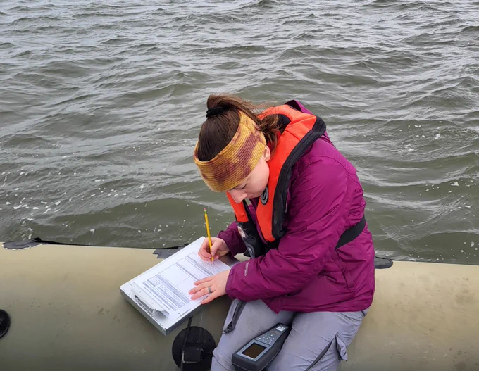 woman sits on side of a boat recording information on a clipboard