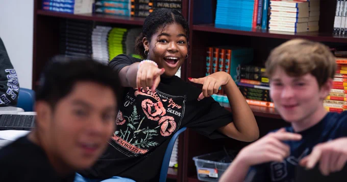 Three students are sitting in a classroom in front of a bookshelf filled with colorful books. The student in the center, a girl wearing a black floral graphic T-shirt, is smiling brightly and pointing toward the camera with both hands. The two boys on either side of her are slightly blurred but also appear to be smiling and gesturing playfully. The mood is upbeat and fun.
