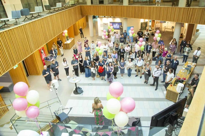 This is a photo of an indoor event where a woman speaks to a crowd gathered in a modern atrium.  Behind her are pink green and white balloons and a sting of bunting and a tv screen.  