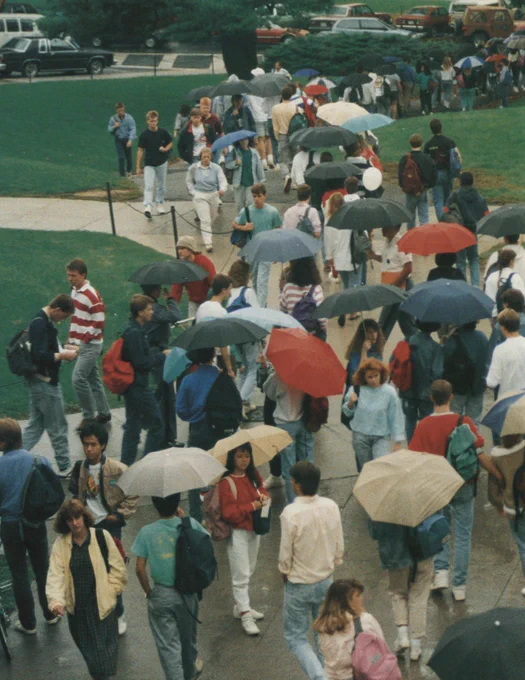 A crowded college campus walkway on a rainy day in the late 1980s or early 1990s. Many students are walking with umbrellas of various colors, wearing casual clothing like jeans, sweatshirts, and backpacks. The fashion includes high-waisted pants, oversized sweaters, and layered looks. The ground is wet, and green lawns surround the paved paths. In the background, vintage cars are parked along the street, adding to the retro atmosphere.


