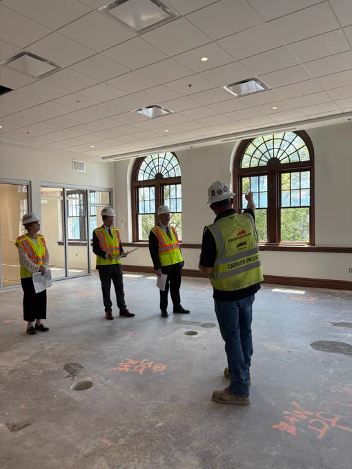 President Jim, wearing a construction hat and vest, is shown a reading room with a view of main campus. 