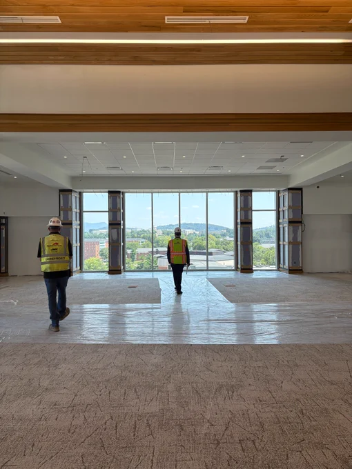 President Jim, wearing a construction hat and vest, walks through a reading room with a view of the mountains and East Campus.
