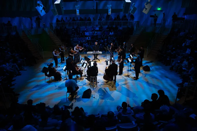 18 musicians in a circle on a blue-lit stage surrounded by audience members, performing “FLOW Symphony." Photo: Jonathan Sachs