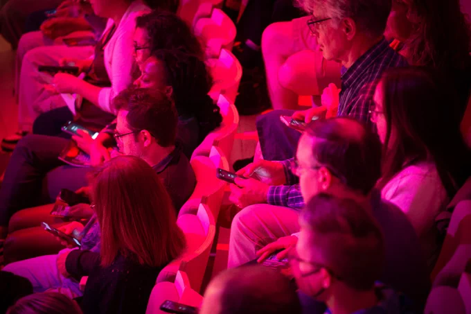 Closeup on about 10 red-lit audience members, all holding cell phones, as part of the performance of “EV6.” Photo: Jonathan Sachs