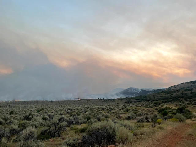 smoke creates clouds of haze in the sky while a field of sagebrush burns on a landscape of fields and mountains