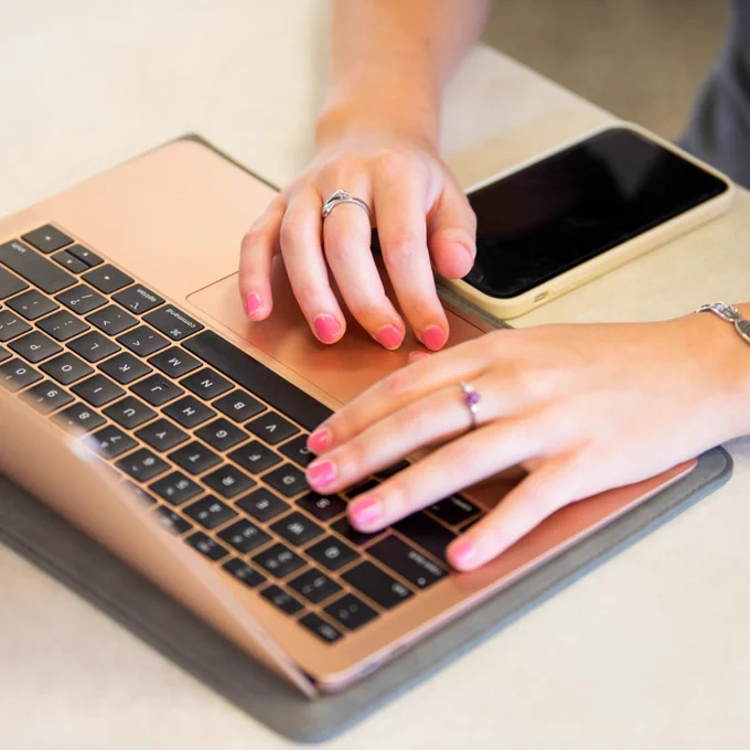 A student with pink nail polish types on a rose gold laptop next to a smartphone and a small stack of library books. 