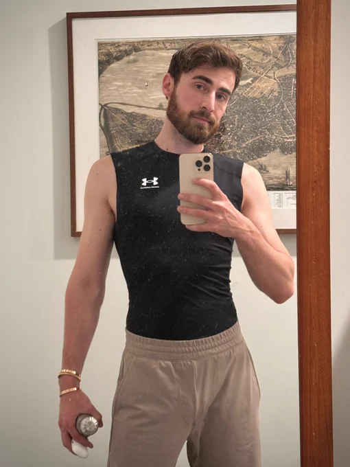 Bearded man taking a selfie in front of a mirror. Wearing black Under Armour compression shirt and tan sweatpants. Holding steel water bottle and phone. 
