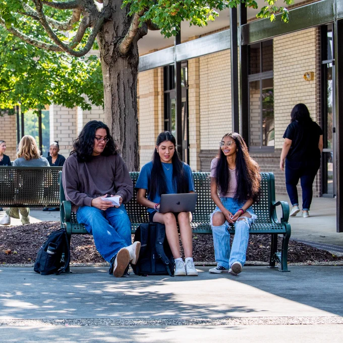 Students gather outdoors on a college campus, sitting on benches and talking under leafy green trees. Three students sit on one bench, with one using a laptop, while others are seated or walking in the background. The scene is bright and welcoming, with sunlight filtering through the trees and brick buildings surrounding the area.