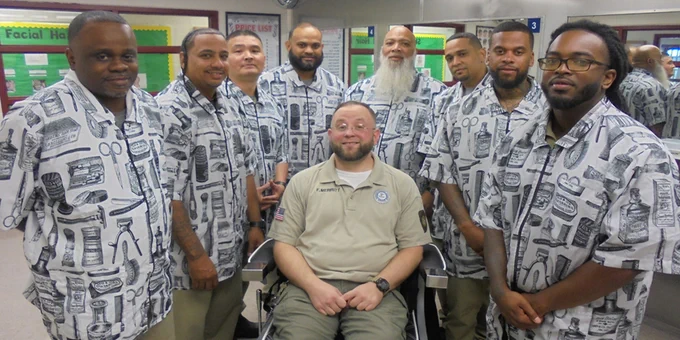 Group of BOP inmate barber school student in matching patterned shirts, smiling in a class with  the staff instructor seated in front in a barber chair.