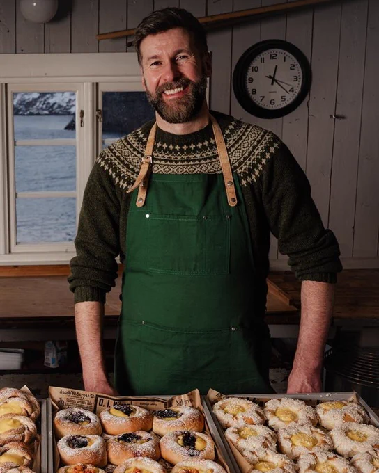 A man wearing a knitted jumper stands and an apron  proudly behind a lot of cakes