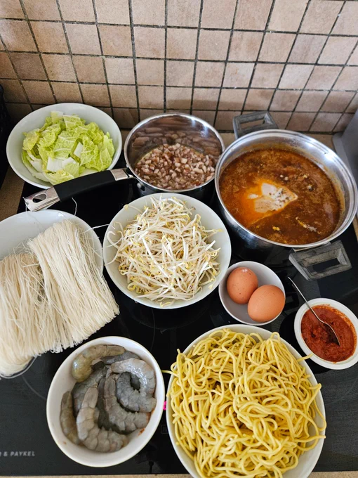 Ingredients clockwise from top left of a bowl of bokchoy pieces, a pot with small cubes on pork fat & skin in oil, a pot of the spicy broth with a piece of pork in, a small bowl with two eggs, a small dish of sambal, a bowl of yellow wheat noodles, a small bowl of uncooked whole prawns, a bowl of rice vermicelli, a bowl of beansprouts.
