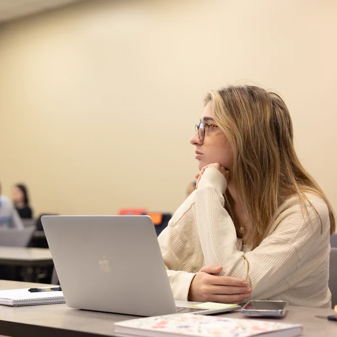 A college classroom with students seated at desks. In the foreground, a young woman wearing glasses and a cream-colored sweater is focused and looking toward the front of the room. She has a laptop, notebook, and phone on her desk. Other students are visible in the background, slightly out of focus.
