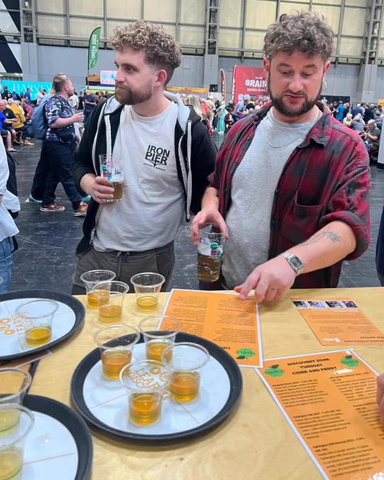 Two young men stood at CAMRA's Great British Beer Festival Discovery Zone bar, sampling ciders. There are three trays of small samples of beer and cider.