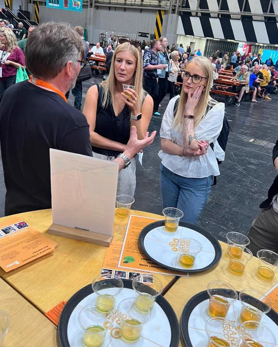 Two young women sampling cider at CAMRA's Great British Beer Festival Discovery Zone while a man is educating them on the cider. There are three trays on the bar filled with small samples of cider and beer.
