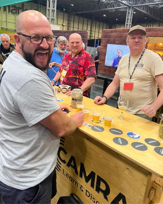A man is stood at CAMRA's Great British Beer Festival Discovery Zone bar, talking to two volunteers behind the bar while sampling cider.