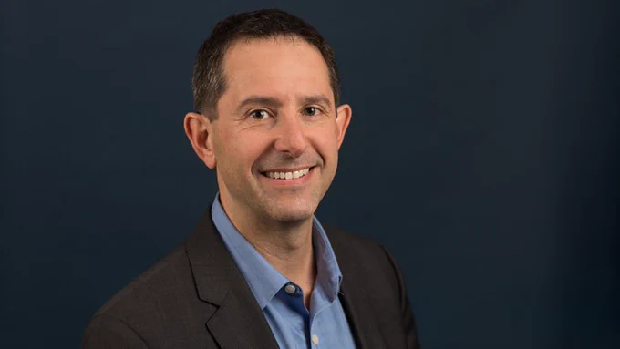 Portrait of Peter Stone, smiling, in a dark suit and light blue shirt against a navy background.