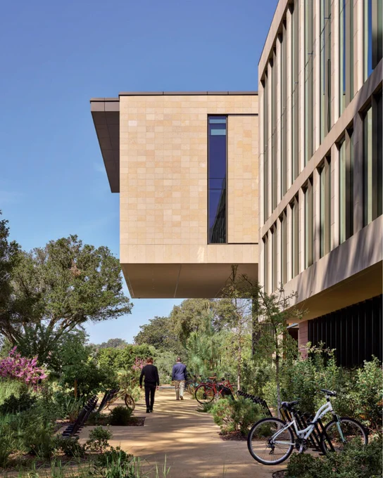 Exterior view  at a university, featuring a modern building with an overhanging section. People are walking on a pathway surrounded by lush greenery and parked bicycles.