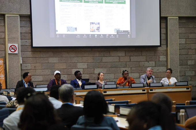 Six panelists seated at a conference table in front of a large screen displaying a presentation, inside a conference hall.