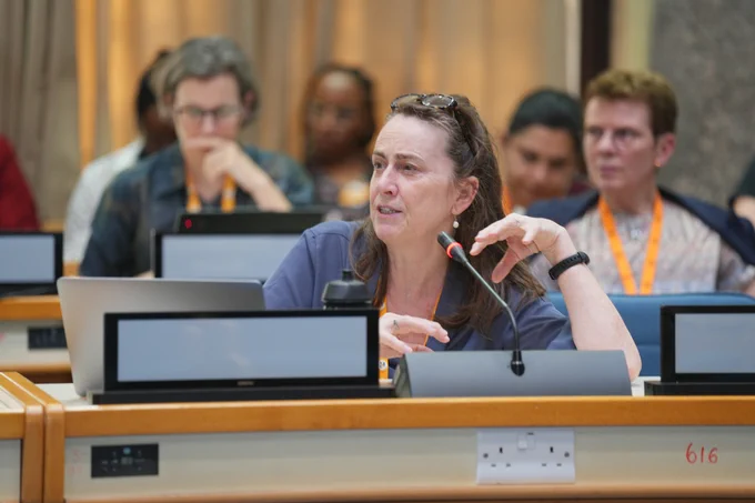 A woman speaking into a microphone attached to a desk during a plenary session at CGIAR Science Week, with attendees and laptops in the background.