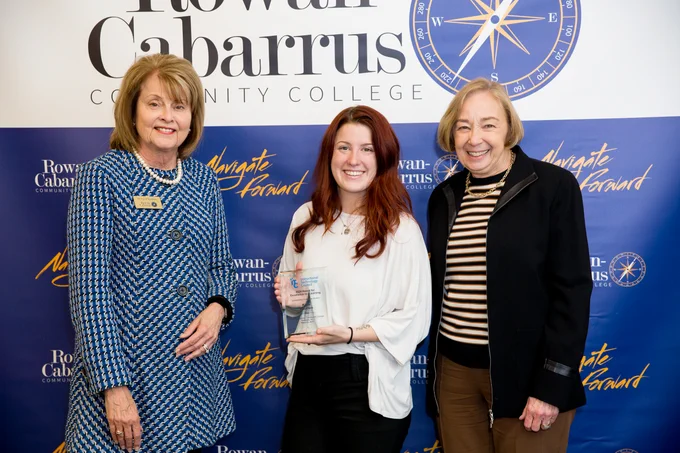 Three women stand in front of a Rowan-Cabarrus Community College backdrop. The woman in the center holds an award and smiles. The women on either side of her are dressed formally and also smiling, marking a celebratory occasion.