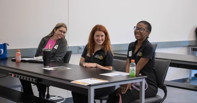 Three women sit at a long table in a classroom or meeting room. All are smiling toward the camera. The woman in the center has red hair and is wearing a black Rowan-Cabarrus Community College polo shirt. Papers, drinks, and personal items are on the table.