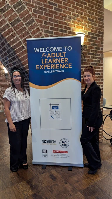 Two women stand on either side of a vertical banner that reads “Welcome to the Adult Learner Experience Gallery Walk.” The event is held in a venue with brick walls and wood flooring. The banner features logos for NC Community Colleges, NC SSC, NC State University, and others.