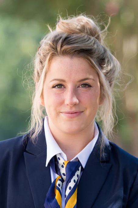 A young woman with blonde hair tied up in a messy bun, wearing a navy blue blazer, white collared shirt, and a navy, yellow, and white polka-dotted scarf. She is standing outdoors with a blurred green background, smiling slightly at the camera.
