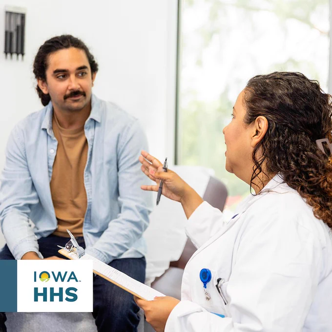 A person in a white lab coat converses with another individual in a denim jacket during a consultation session, with an "Iowa HHS" logo visible in the corner of the image.