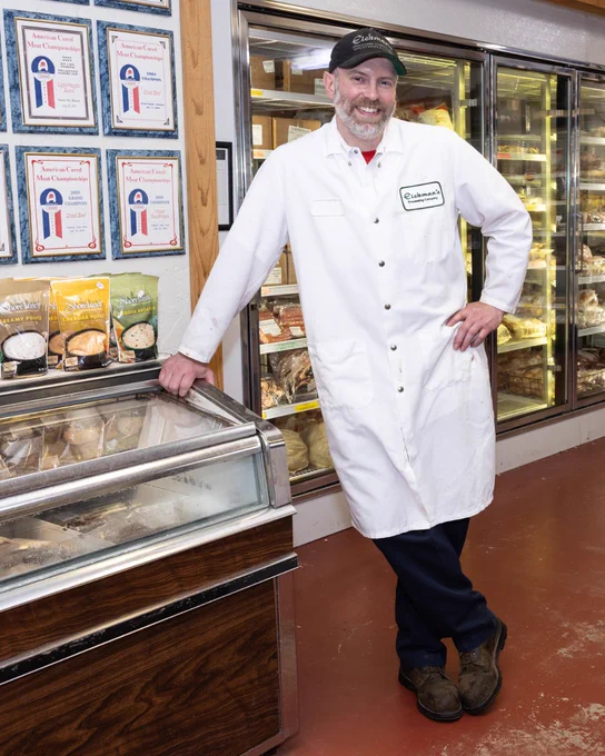 An individual in a white coat stands smiling in a grocery store beside a freezer filled with packaged food products.