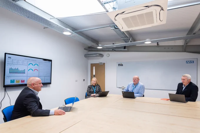 Four professionals seated around a large conference table with laptops during a meeting in a modern office room.