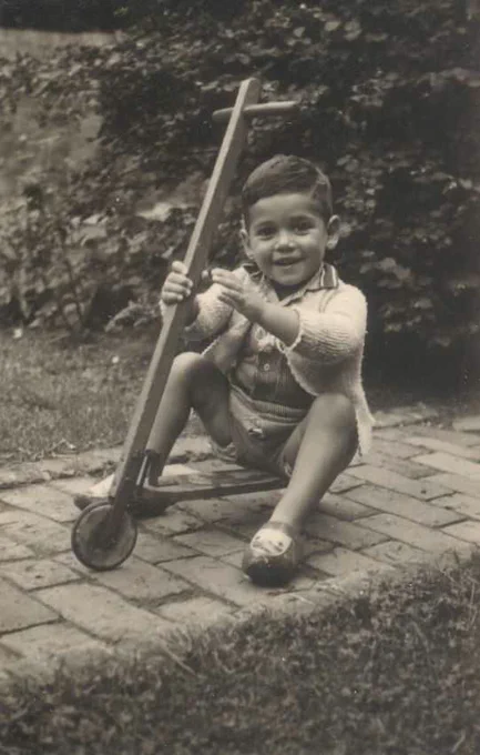 A vintage black and white photo of a young childboy smiling and sitting on a brick pathway, on a scooter. The background features a garden with visible foliage.
