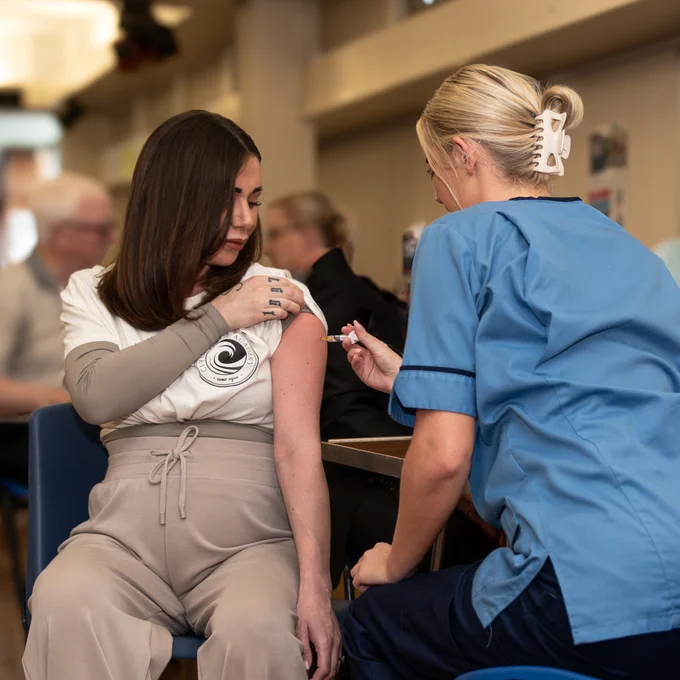 A pregnant woman sitting and pulling up her sleeve while a nurse administers the RSV vaccine in her arm.