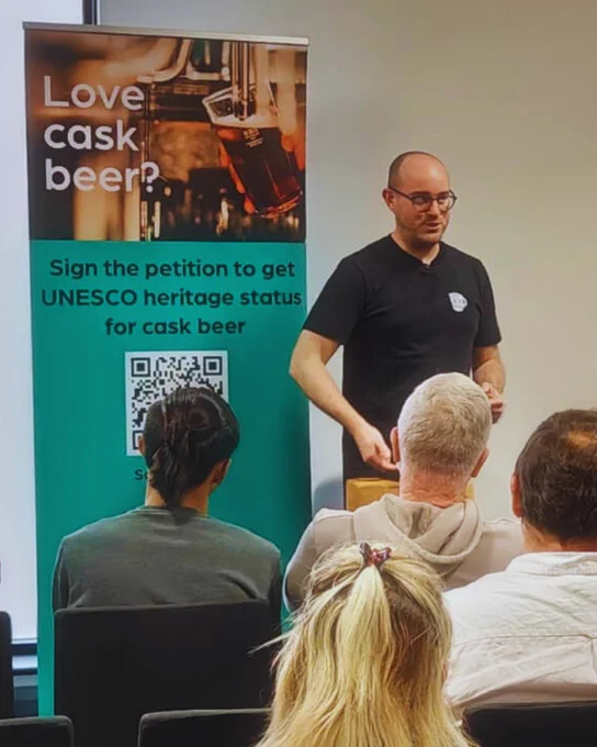 Jonny Garrett is standing next to a green roller banner that has a QR code linking to the Parliamentary petition to get cask beer UNESCO heritage status. Black text above the QR code that says Sign the petition to get UNESCO heritage status for cask beer. He is talking to a group of people at his panel at Great British Beer Festival.