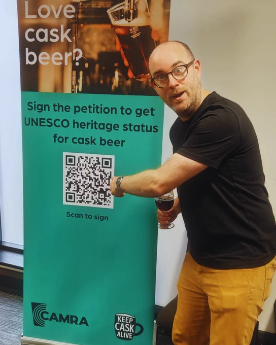 Jonny Garrett is pointing at a green roller banner that has a QR code linking to the Parliamentary petition to get cask beer UNESCO heritage status. Black text above the QR code that says Sign the petition to get UNESCO heritage status for cask beer.