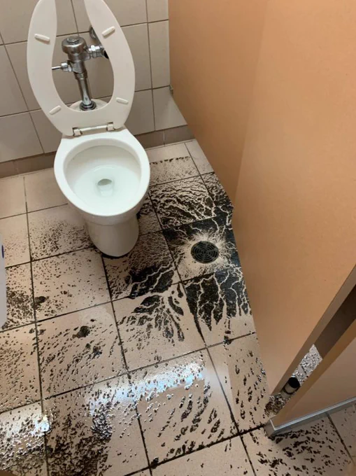 A washroom stall with a dirty floor covered in sand and an open toilet.