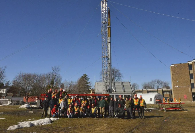 A group of students pose in front of a tall rocket on a launch pad, under a clear blue sky.
