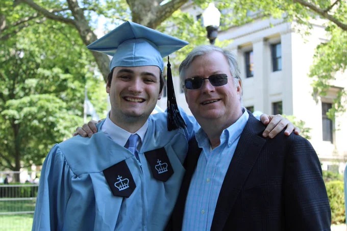 Evan Rork '23GS in a Columbia graduate gown on Columbia's campus, posing, smiling, with a friend/family member