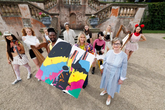 Group of people of various ages posing with a colorful abstract painting, some in costumes and holding musical instruments, at an outdoor location with ornate architectural details in the background.