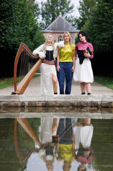 Three people pose outdoors, one holding a harp, at the edge of a reflecting pond with trees and a building in the background.