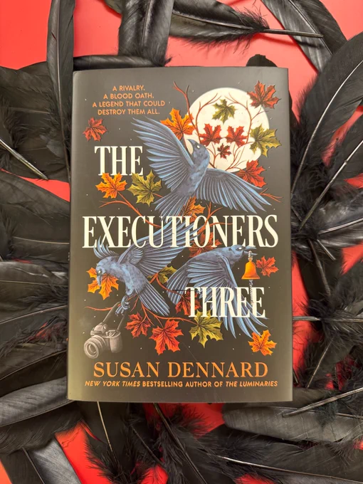 A copy of The Executioners Three by Susan Dennard resting on a red background covered in black feathers.