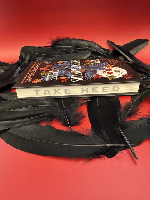 A copy of The Executioners Three by Susan Dennard resting on a red background covered in black feathers with the edges facing the camera. On the edges are the words: "Take Heed."