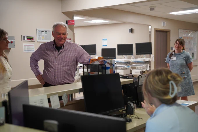 Senator Kaine speaks to a woman sitting at a desk with a computer. 