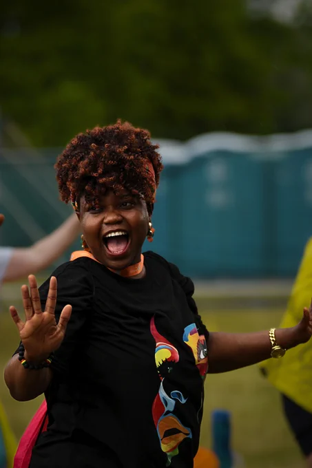 A photo of a lady with her hair up and wearing a black t shirt with an illustration of a lady on it in block colours. She is mid dance with her hands out on either side and a huge open smile on her face!  