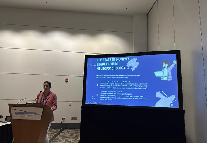 A woman standing behind a podium, with a presentation behind her with a slide saying 'the state of women's leadership in neuropsychology.'